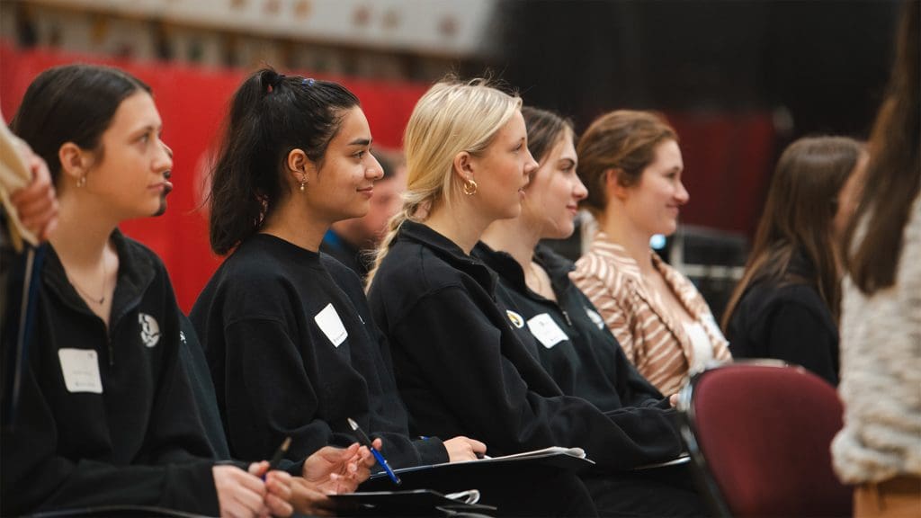 a group of high school women are seated in a row facing to the right smiling while listening to a presentation